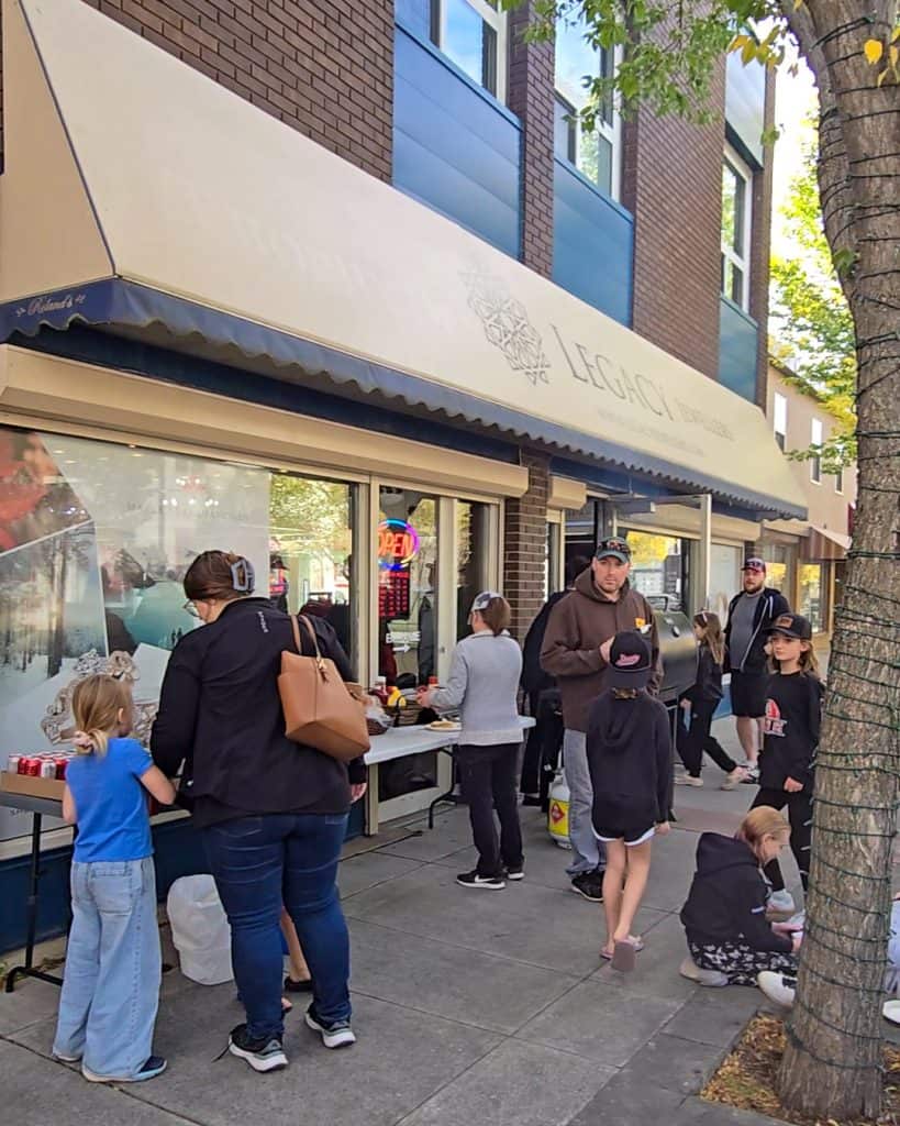 BBQ setup with families lining up for food, enjoying the sunny weather and good company outside the store.